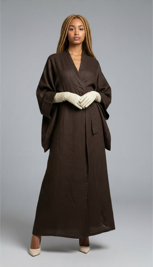 A model wearing a dark brown mud dyed linen wrap-style maxi robe with wide draped sleeves, paired with cream gloves and beige heels, standing against a neutral gray studio background.