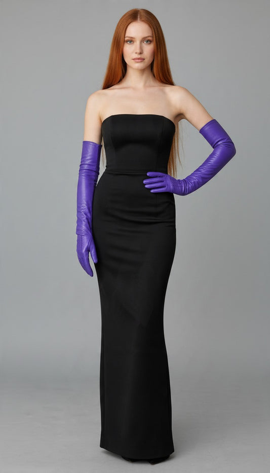 A woman modeling a strapless black floor-length gown with long purple opera gloves against a plain gray studio background.